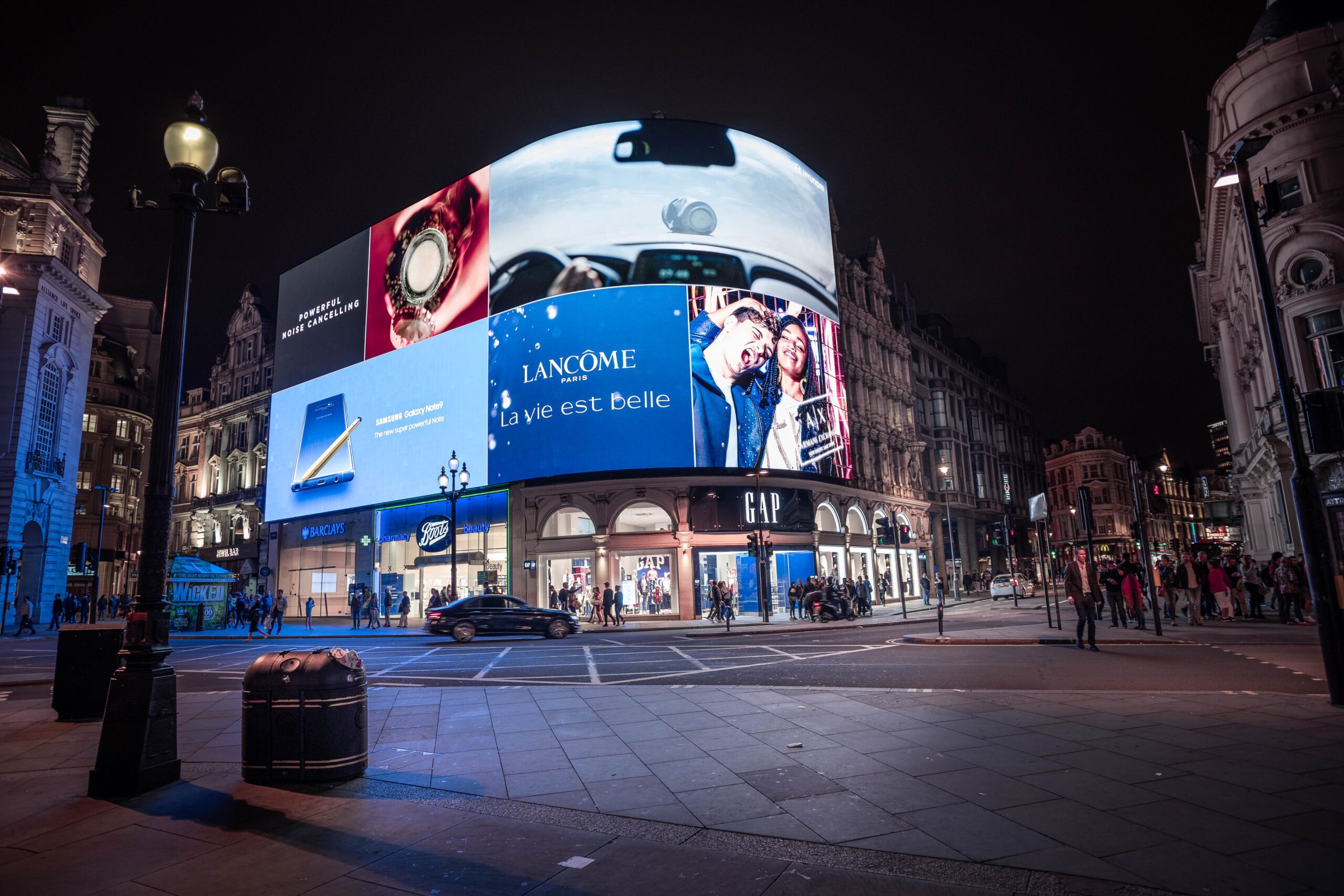 A vibrant urban scene at night showcasing smart digital signage on a large curved LED display. The screen displays high-resolution advertisements, including luxury brands and tech products, demonstrating the impact of dynamic, energy-efficient digital signage in engaging audiences in a busy commercial area. The scene highlights the power of smart signage for marketing in city environments.