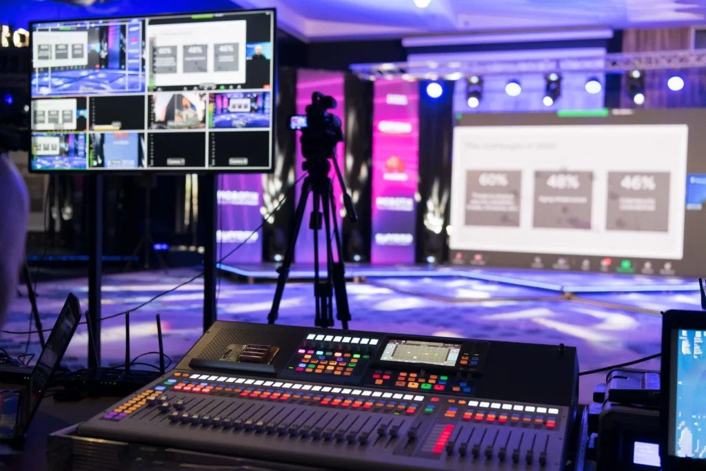A professional event production setup showcasing immersive event technology solutions. The image features a colorful stage illuminated with purple and blue lighting, a large digital screen displaying presentation content, a live video feed on a monitor, and an advanced audio-visual control console in the foreground, emphasizing the seamless integration of technology in modern events.