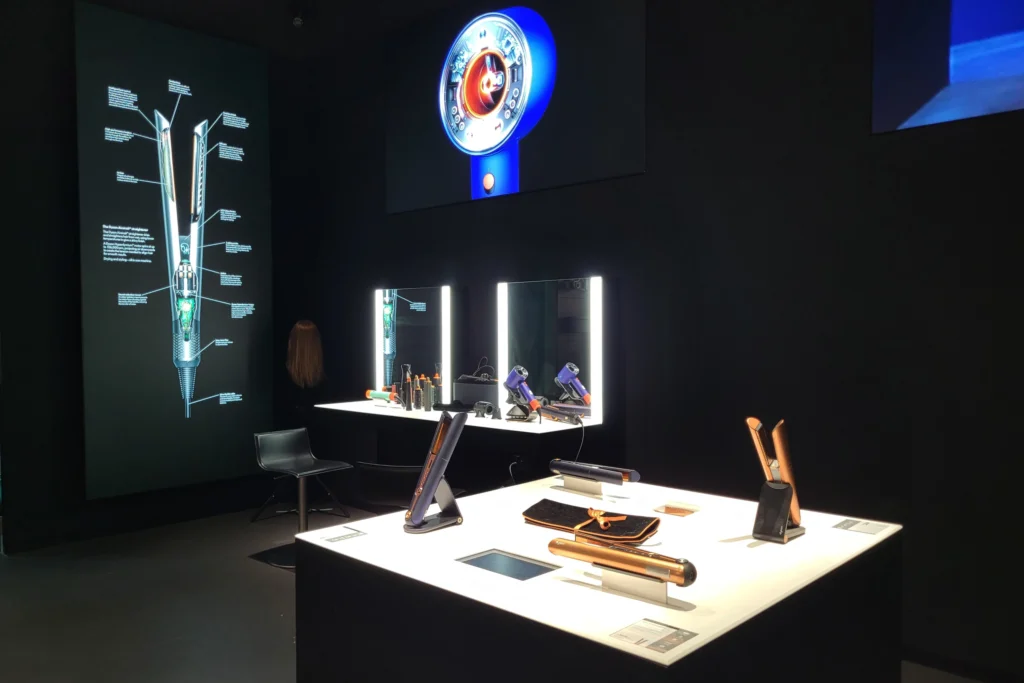 A sleek and modern interactive showroom featuring high-tech beauty tools displayed on illuminated tables. Large digital screens showcase product breakdowns and demonstrations, enhancing the customer journey with hands-on digital experiences. The showroom integrates personalized touchpoints, mirrors for live testing, and immersive visuals, creating an engaging environment that blends physical and digital retail strategies.