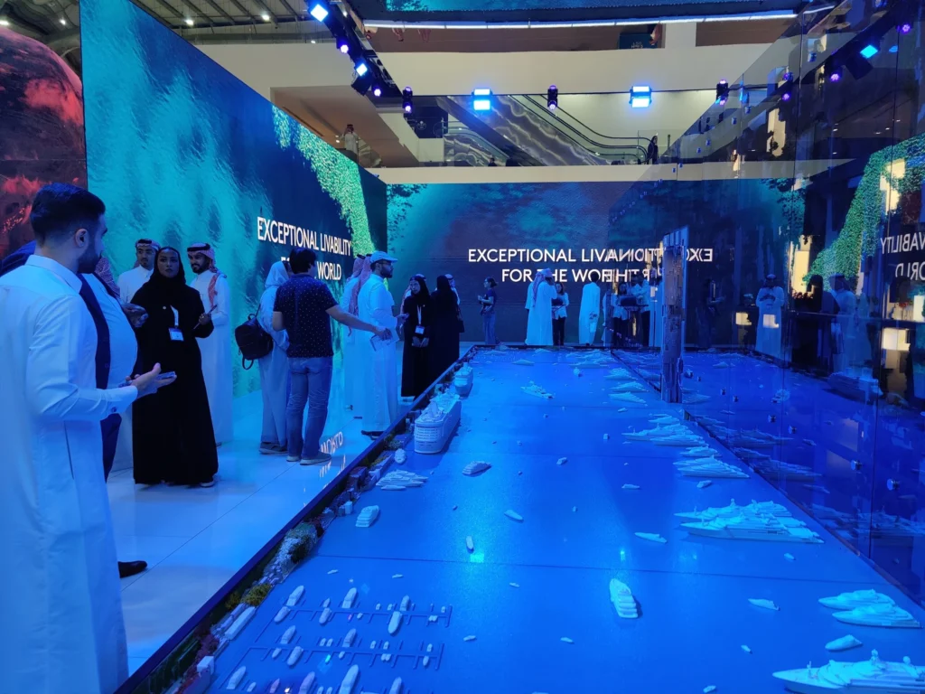 An immersive exhibition showcasing a digital interactive experience, featuring a dynamic floor display of a futuristic cityscape with detailed models and projections. Visitors engage with the vibrant, high-tech environment, enhanced by large LED walls displaying bold visuals and messages, creating a captivating and innovative presentation space.