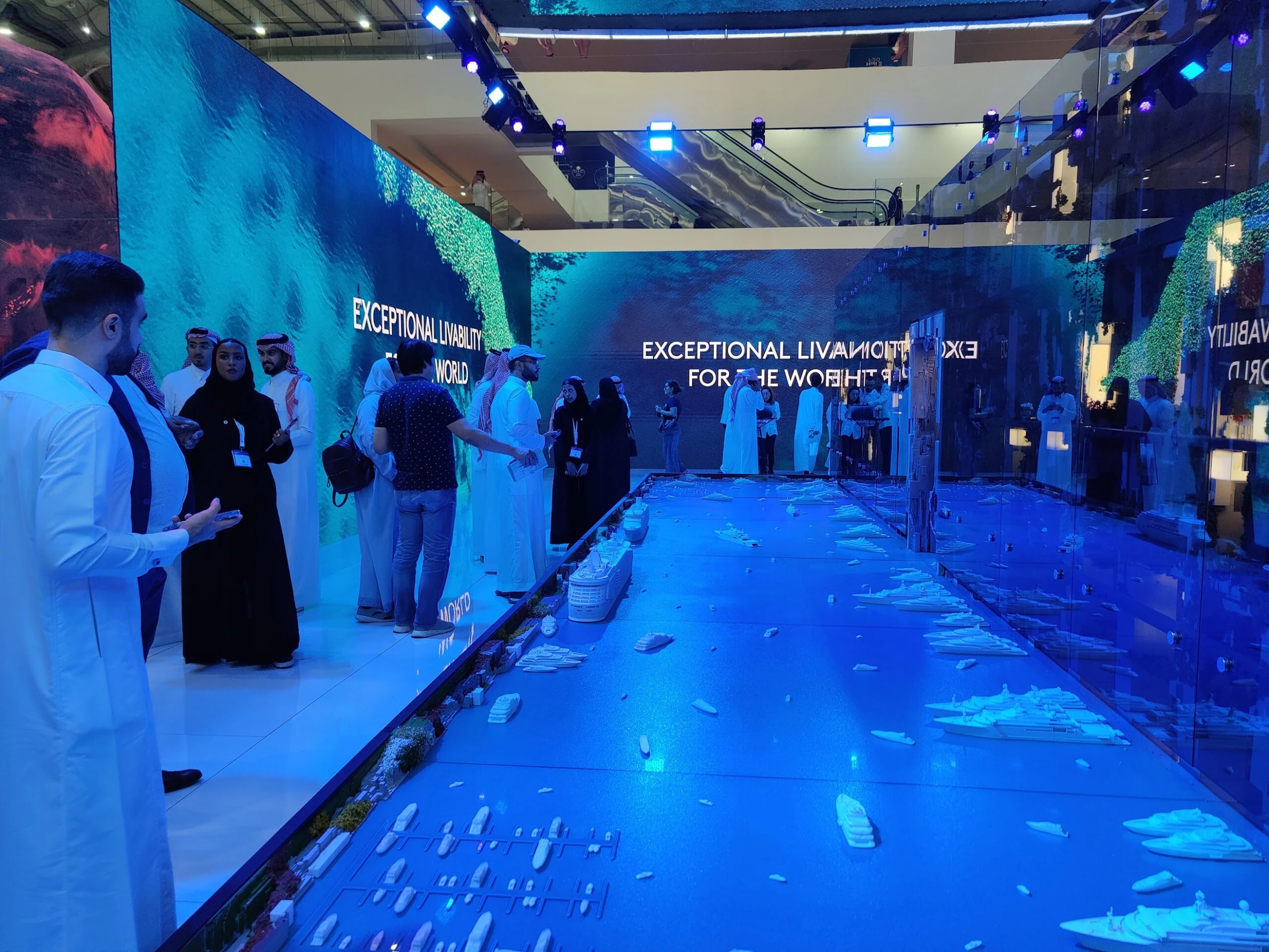 An immersive exhibition showcasing a digital interactive experience, featuring a dynamic floor display of a futuristic cityscape with detailed models and projections. Visitors engage with the vibrant, high-tech environment, enhanced by large LED walls displaying bold visuals and messages, creating a captivating and innovative presentation space.