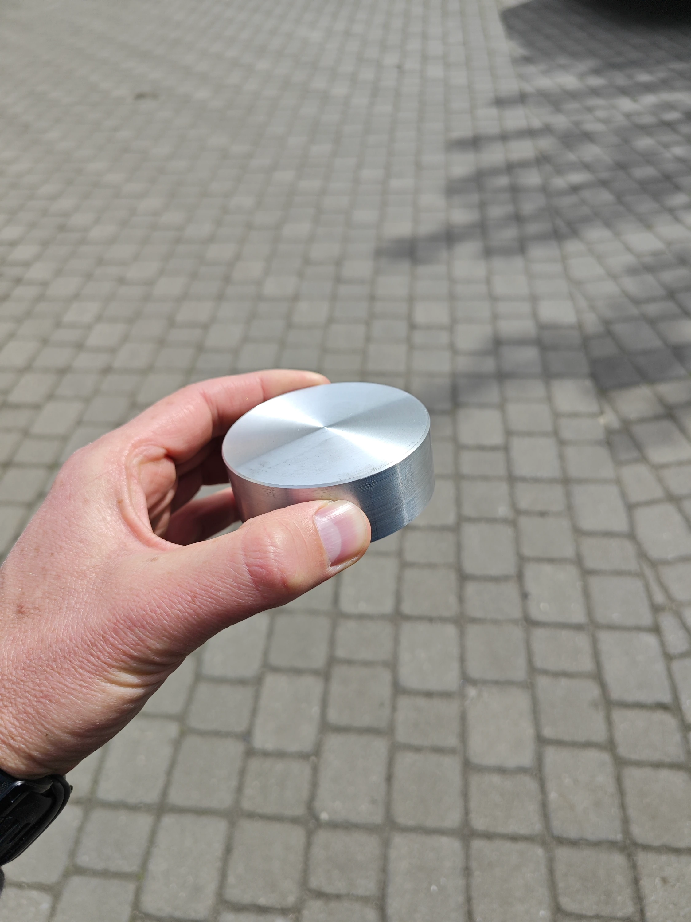 Close-up of a hand holding a freshly milled and polished aluminium token outdoors, showing the satin-finished top surface that will interact with the table.