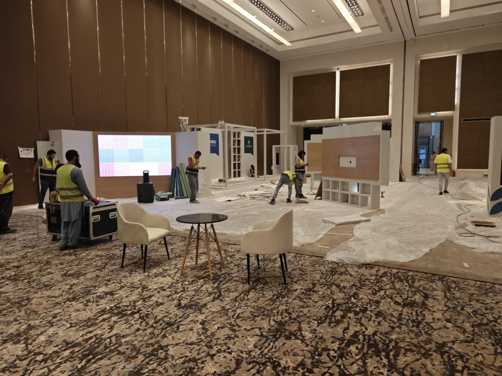 Exhibition hall under construction at AECD 2025: crew in yellow safety vests lay flooring and assemble display units while the interactive table area waits for installation.