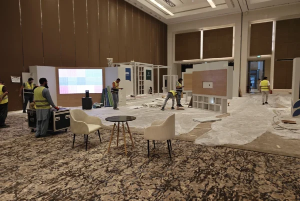 Exhibition hall under construction at AECD 2025: crew in yellow safety vests lay flooring and assemble display units while the interactive table area waits for installation.