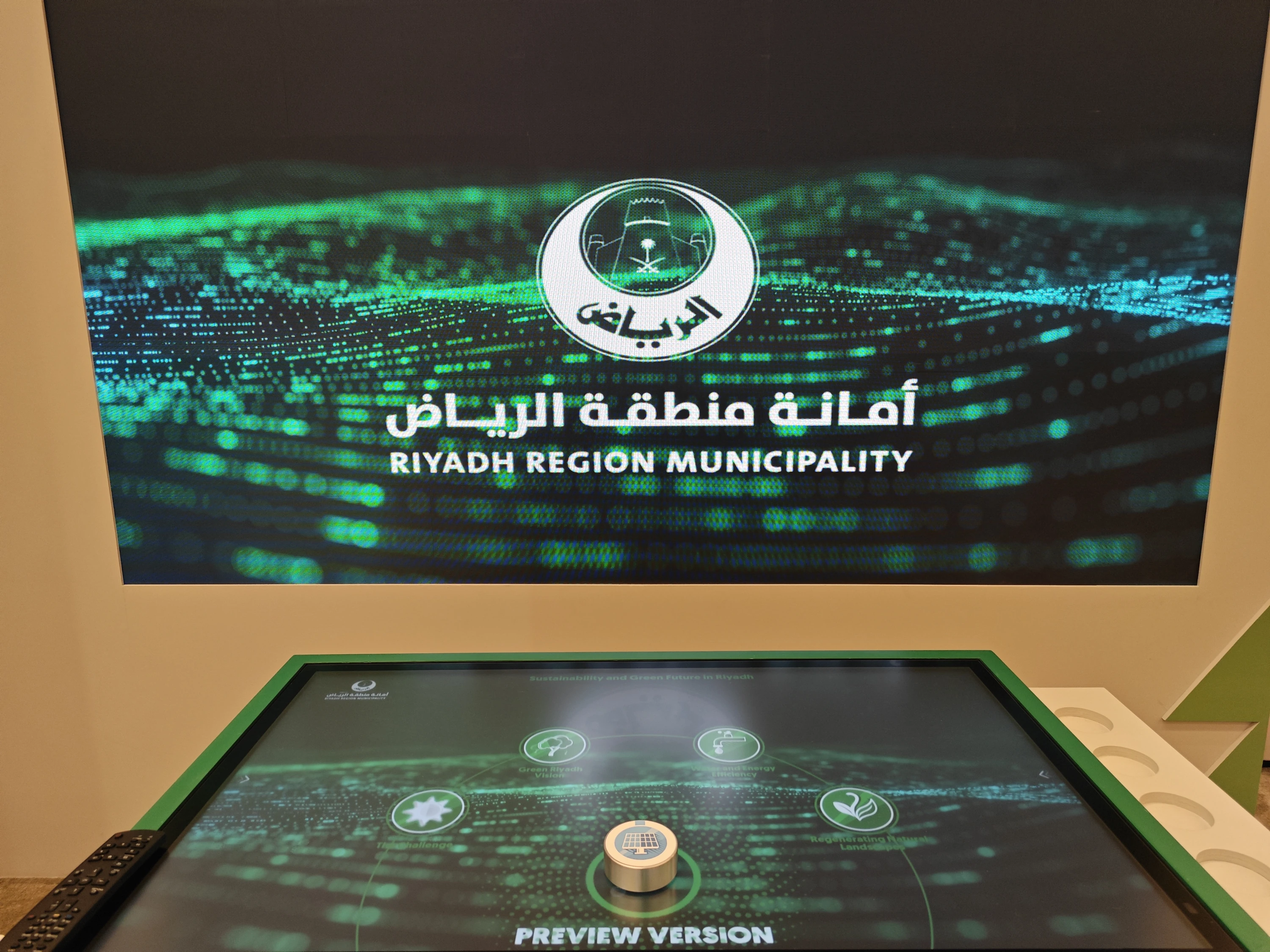 Interactive table powered on during final system checks—Riyadh Region Municipality logo animates on a large LED wall behind the touch surface, with a single aluminium token centred for calibration.