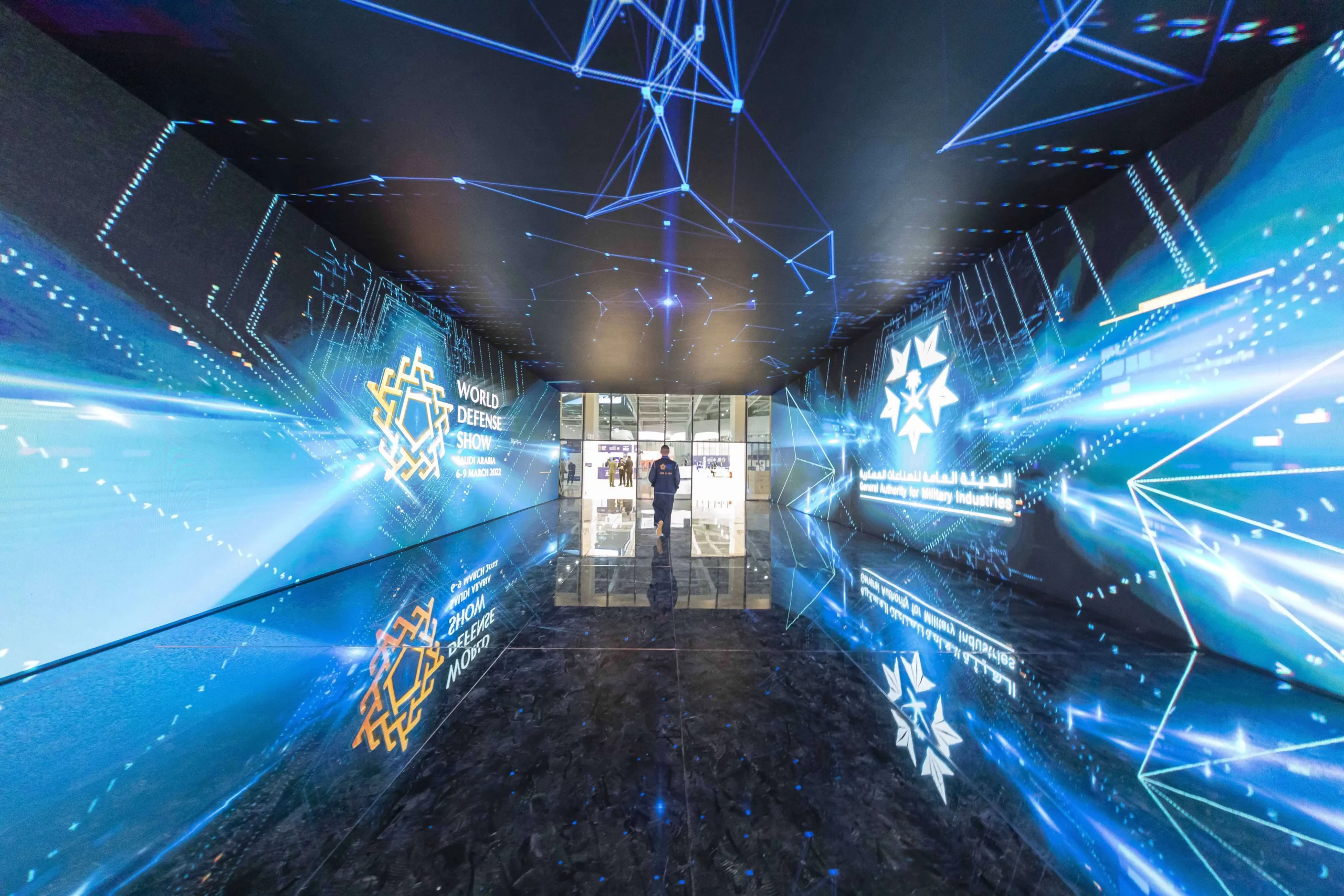 Immersive Booth tunnel at the World Defense Show with floor-to-ceiling LED walls and ceiling displaying dynamic blue holographic graphics as a visitor walks through, reflecting digital patterns on the glossy floor.