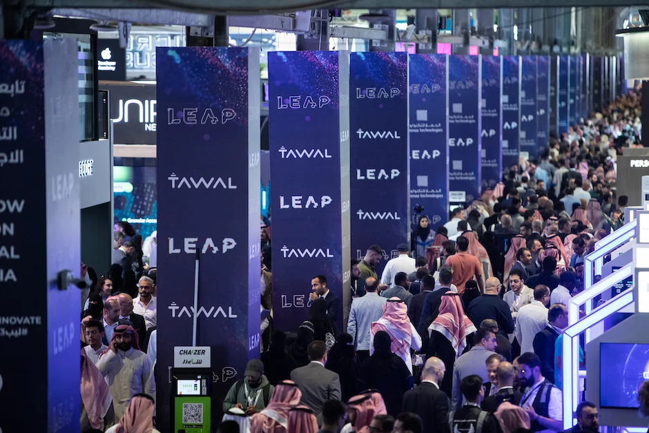 Packed aisle at LEAP tech festival in Riyadh, showing long rows of purple-blue LEAP banners and a diverse crowd of visitors and exhibitors.