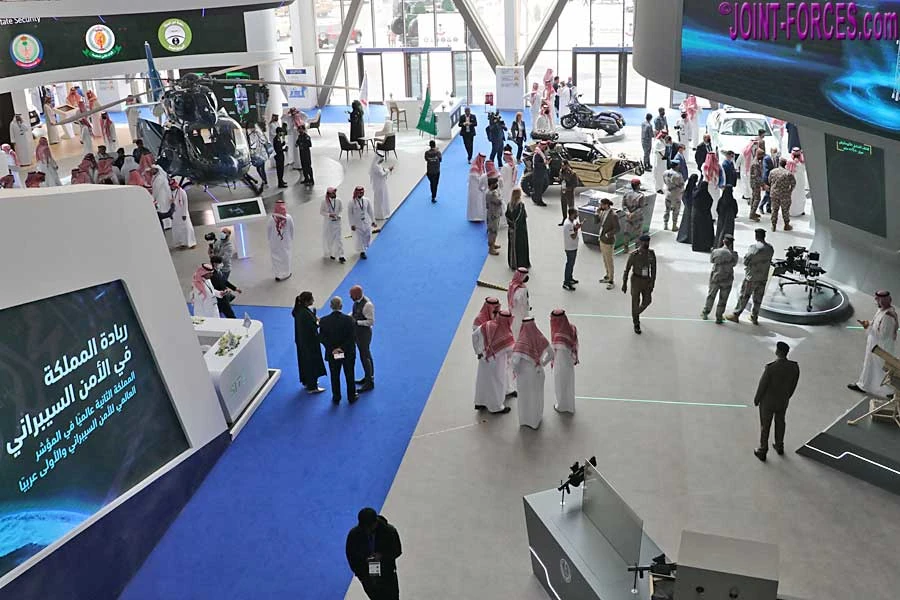Overview of World Defense Show hall in Riyadh: blue carpet walkway, helicopter display, military vehicles, and uniformed as well as civilian delegates.