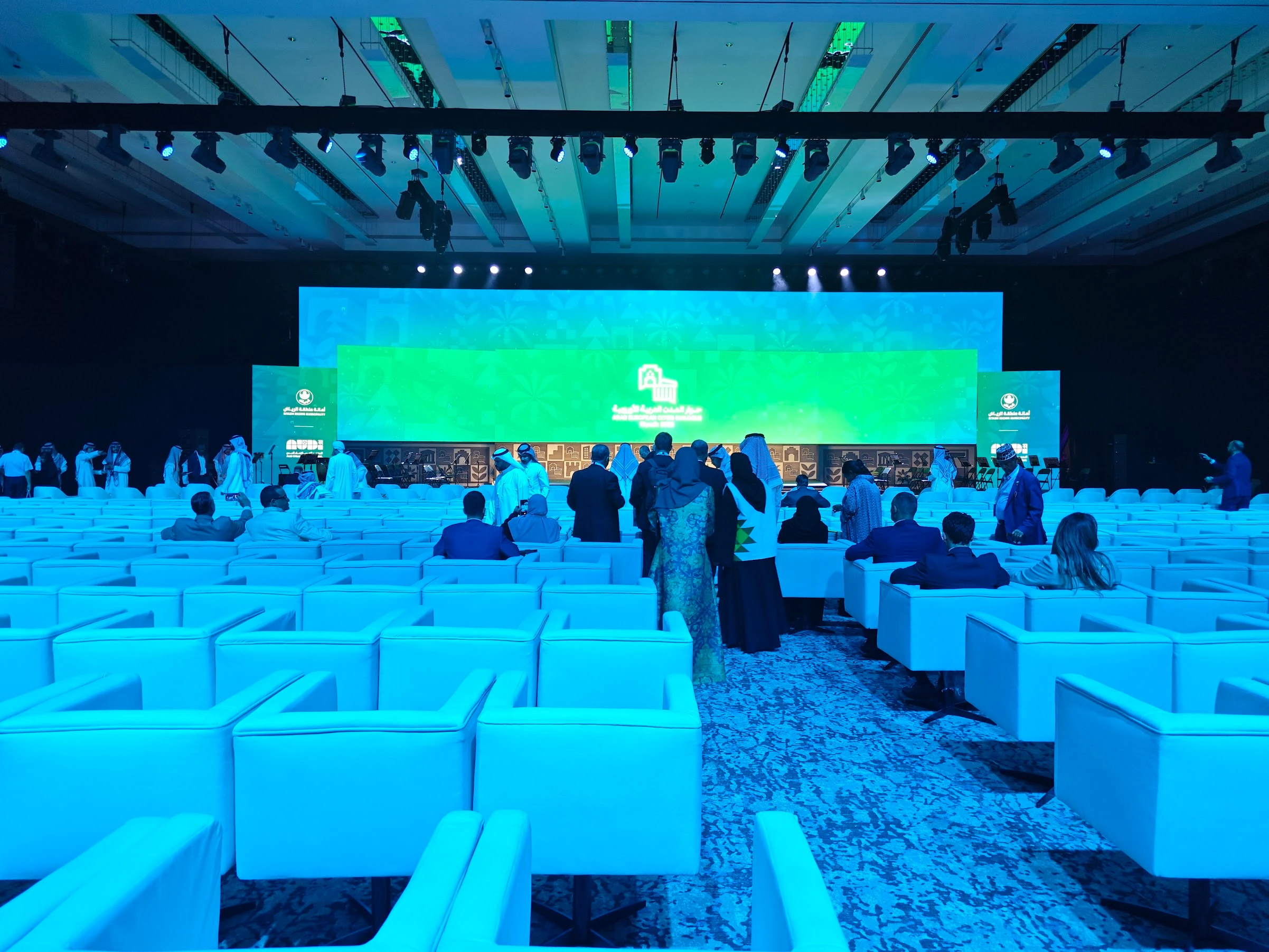 Formal guests mingle among white armchairs facing a wide green LED wall at a high-profile conference—an example of VIP event solutions for corporate summits.