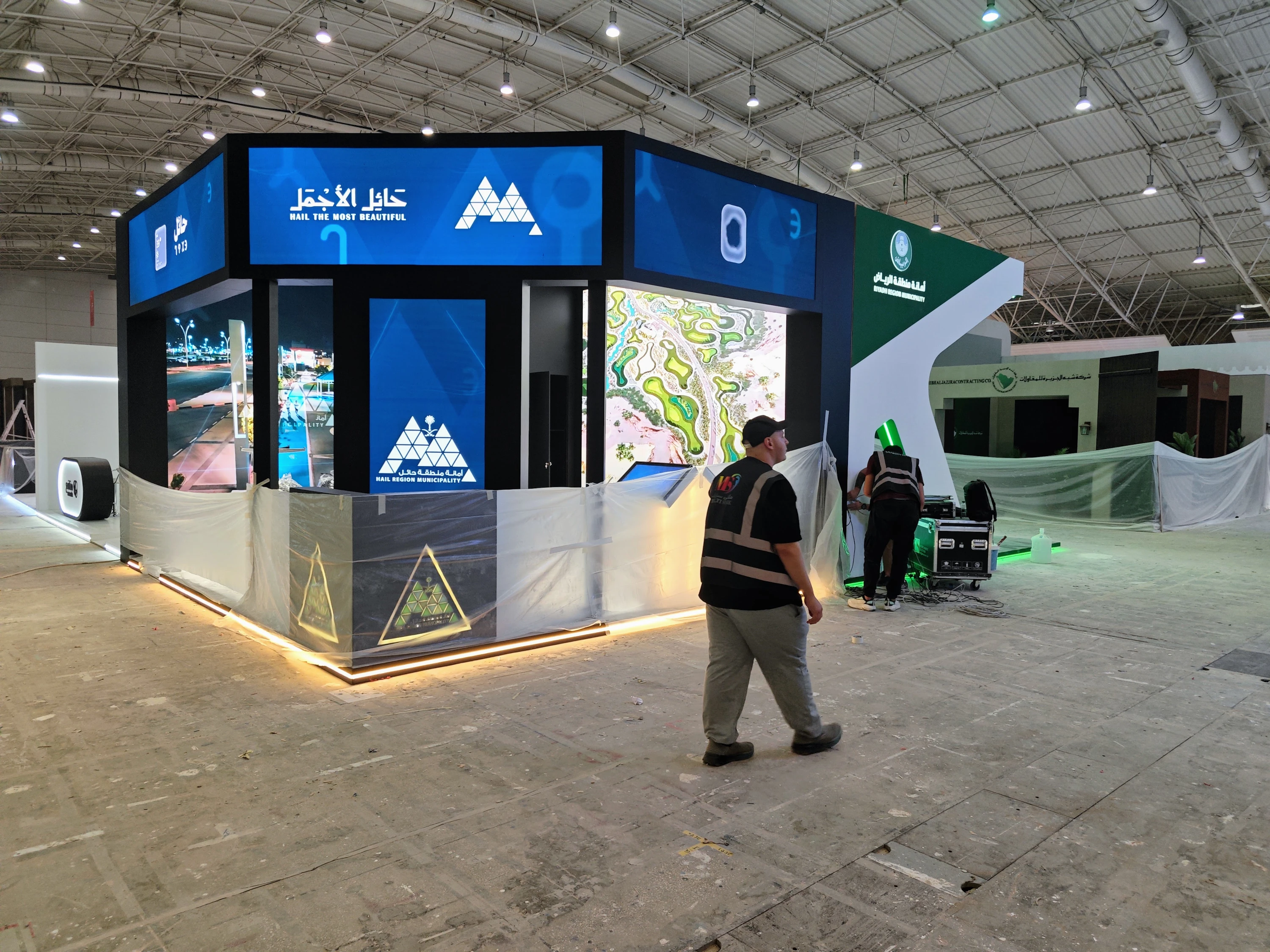 Technicians wiring media servers and LED power as the hall is prepared—neighboring municipal stands being readied alongside the Riyadh Digital Twin at FPF.